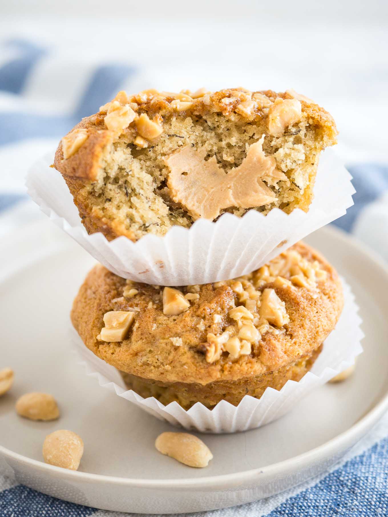 2 muffins à la banane et au beurre de cacahuète, garnis de cacahuètes, avec leurs caissettes ouvertes empilés l'un sur l'autre sur une assiette grise sur un torchon blanc et bleu. Une grande bouchée a été prise dans le muffin du dessus, révélant un cœur au beurre de cacahuète.