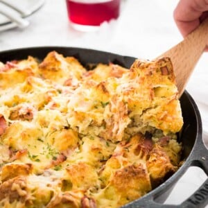 Une poêle en fonte avec un gratin de jambon et de fromage pour le petit-déjeuner. Une main y plonge avec une spatule. En arrière-plan, il y a une pile d'assiettes blanches avec des couverts, un torchon blanc et un verre de thé fruité.