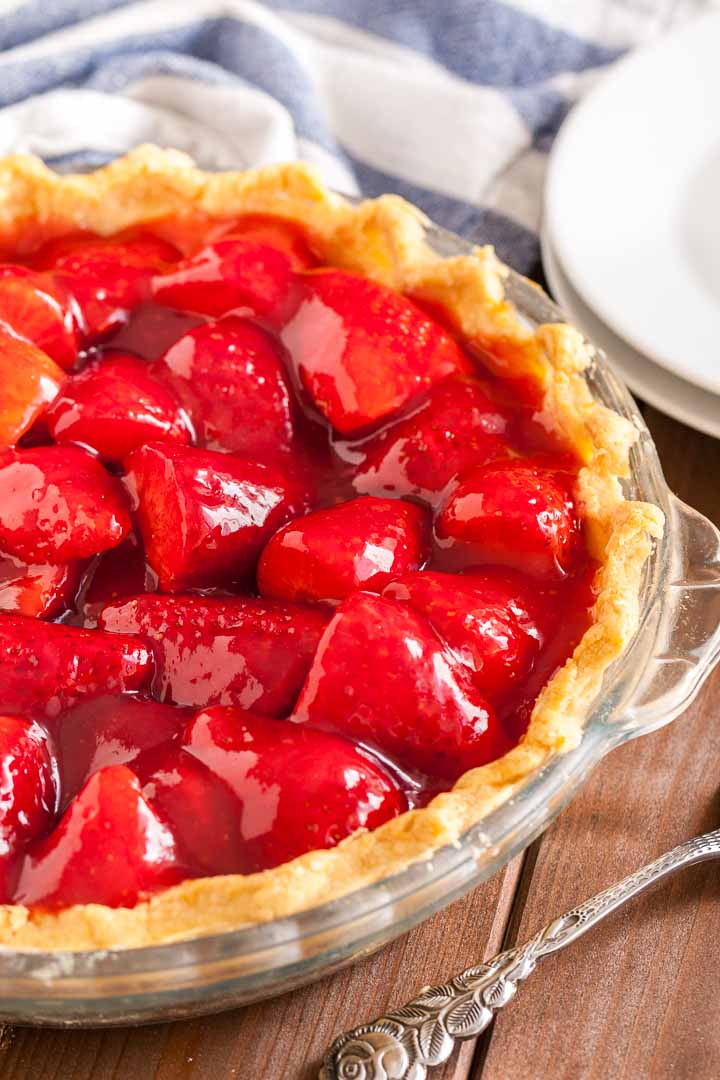 Gros plan d'un plat en verre, avec de la tarte aux fraises sur une table en bois brun, un torchon blanc et bleu, 2 assiettes blanches empilées et un serveur à gâteau.