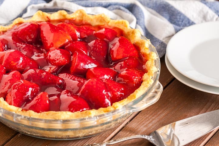 Un plat en verre, avec de la tarte aux fraises sur une table en bois brun, un torchon blanc et bleu, 2 assiettes blanches empilées et un serveur à gâteau.