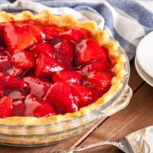 Un plat en verre, avec de la tarte aux fraises sur une table en bois brun, un torchon blanc et bleu, 2 assiettes blanches empilées et un serveur à gâteau.