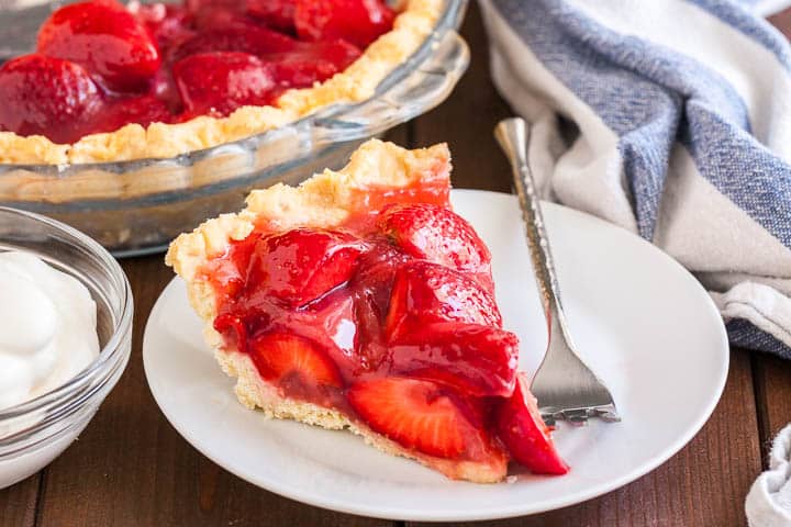 Une part de tarte aux fraises sur une assiette blanche avec une fourchette en argent sur une table en bois. En arrière-plan, il y a un plat en verre avec de la tarte aux fraises et un torchon blanc et bleu.