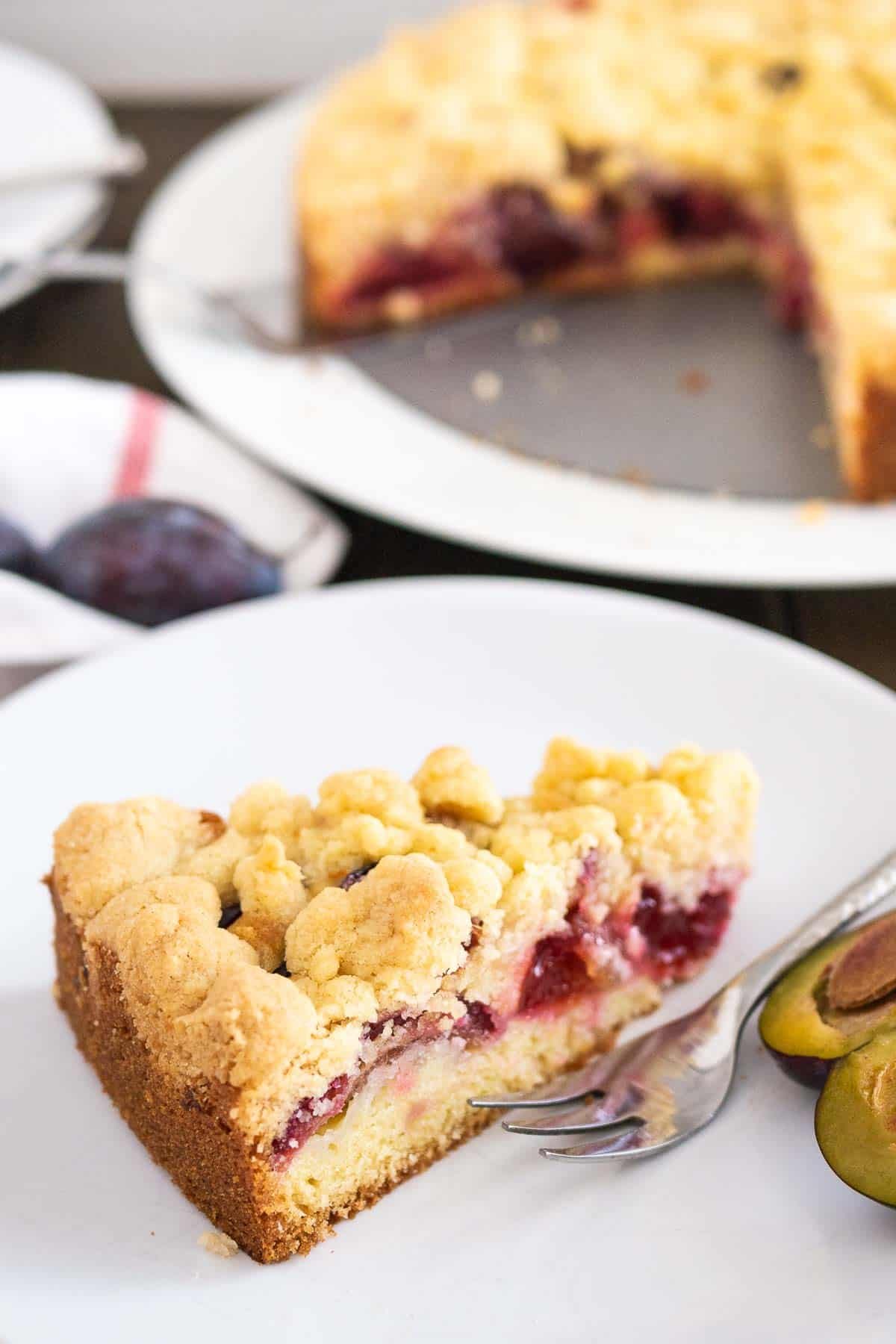 Une part de gâteau aux prunes surmontée de streusel dans une assiette blanche avec une fourchette et une prune coupée en deux. L'ensemble du gâteau est en arrière-plan.