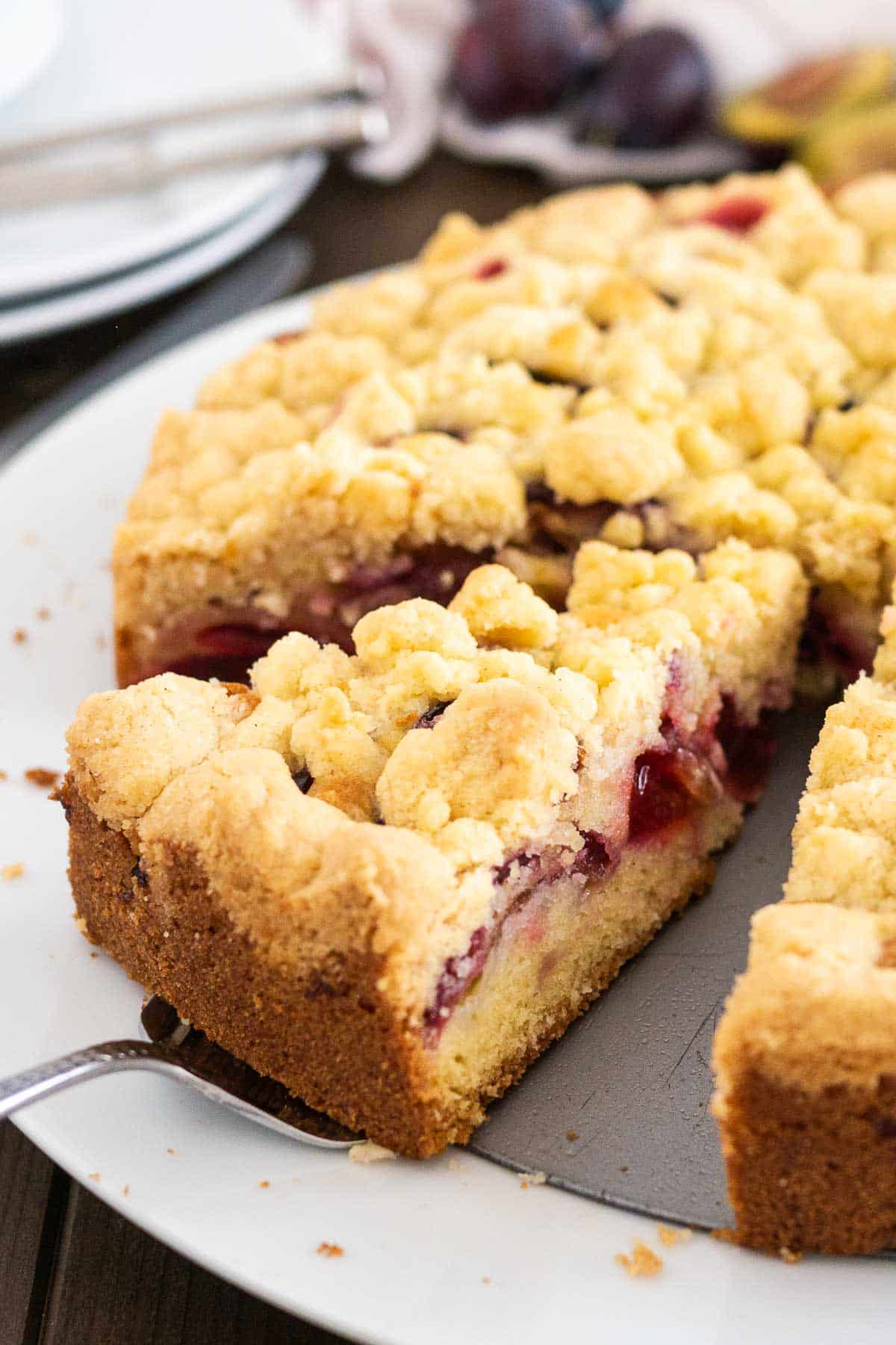 Un gâteau aux prunes avec streusel sur un fond de moule à charnière dans une assiette blanche. Un serveur à gâteau soulève une part.