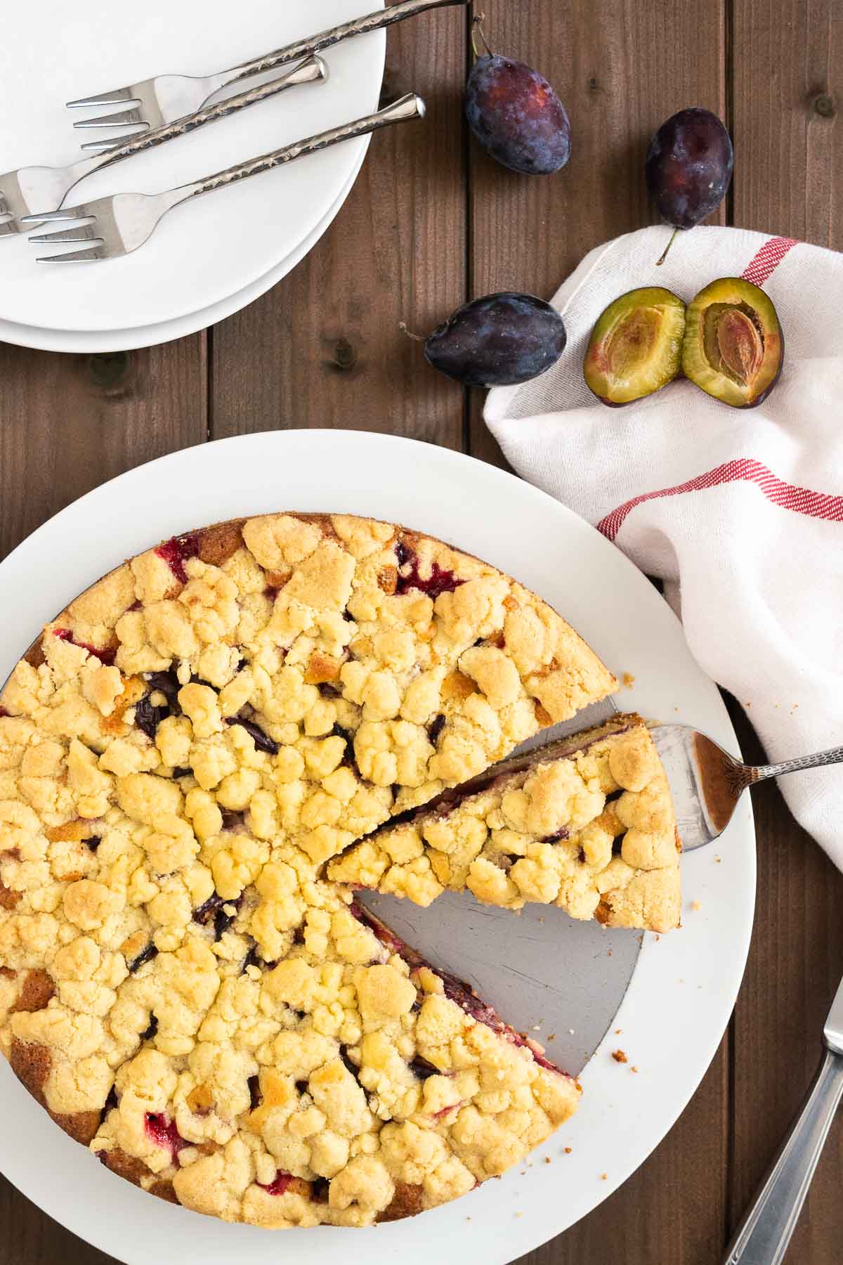 Vue de haut d'un gâteau aux prunes avec streusel sur un fond de moule à charnière dans une assiette blanche. Un serveur à gâteau soulève une part. À côté de l'assiette, il y a des prunes, un couteau, deux assiettes blanches avec des fourchettes et un torchon blanc avec une rayure rouge.
