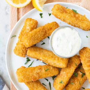 Bâtonnets de poisson sur une assiette blanche garnie de persil sur une planche en bois.