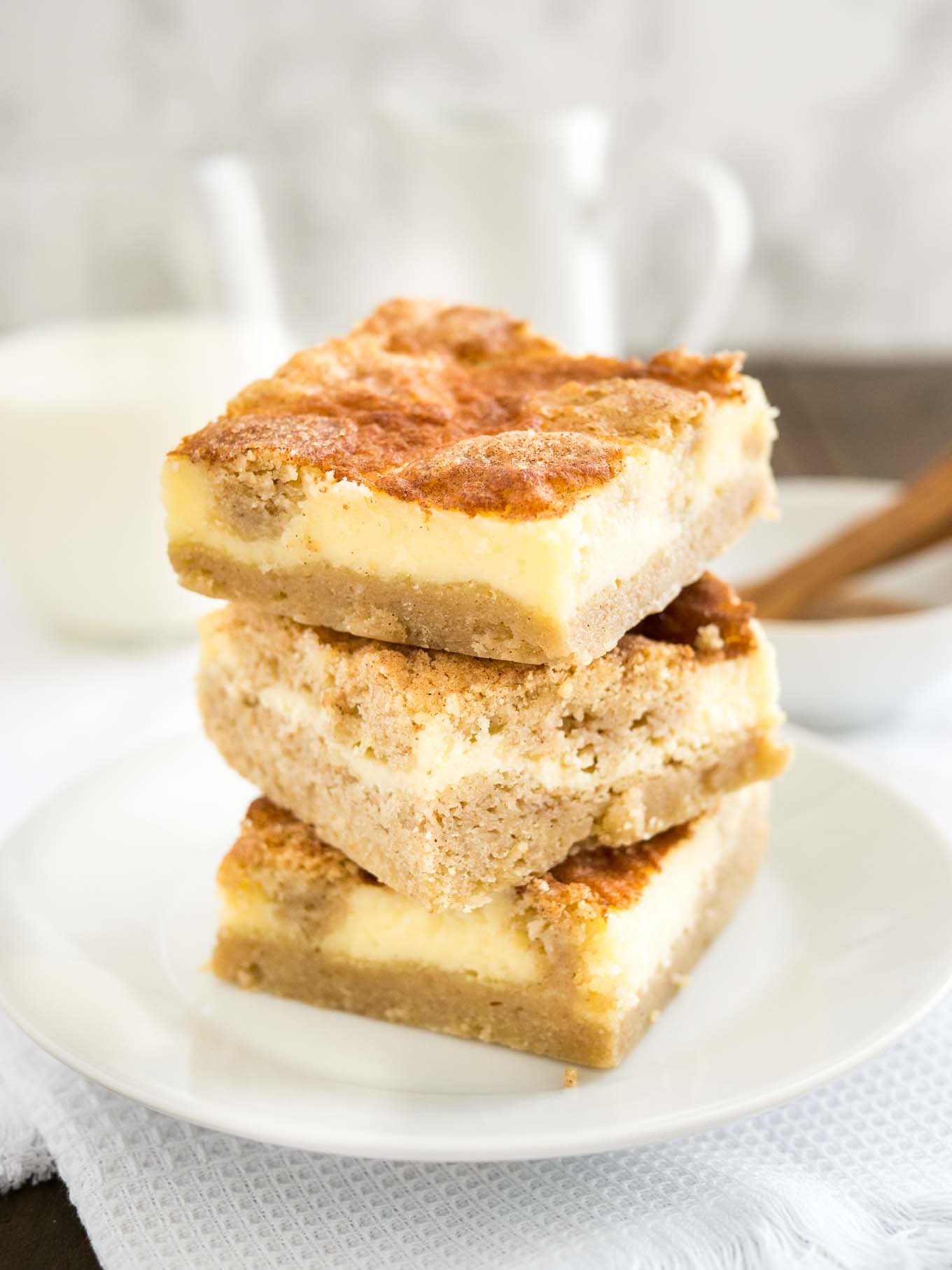 Une pile de 3 barres de cheesecake snickerdoodle sur une assiette blanche sur une nappe blanche. Il y a un bol blanc avec de la cannelle et une cuillère à mesurer ainsi qu'un verre de lait en arrière-plan.