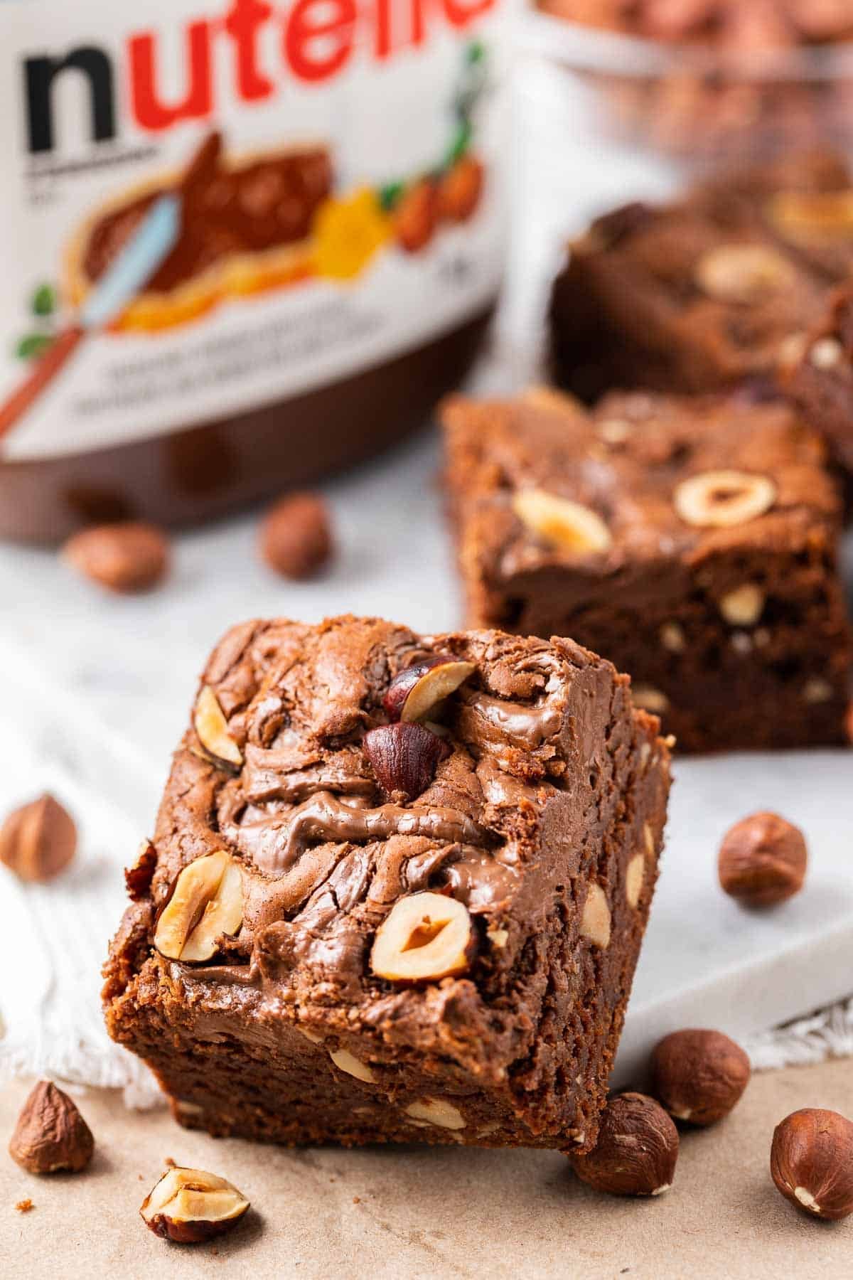Brownie au Nutella s'appuyant contre une assiette blanche avec des noisettes, un pot de Nutella et plusieurs brownies au Nutella en arrière-plan