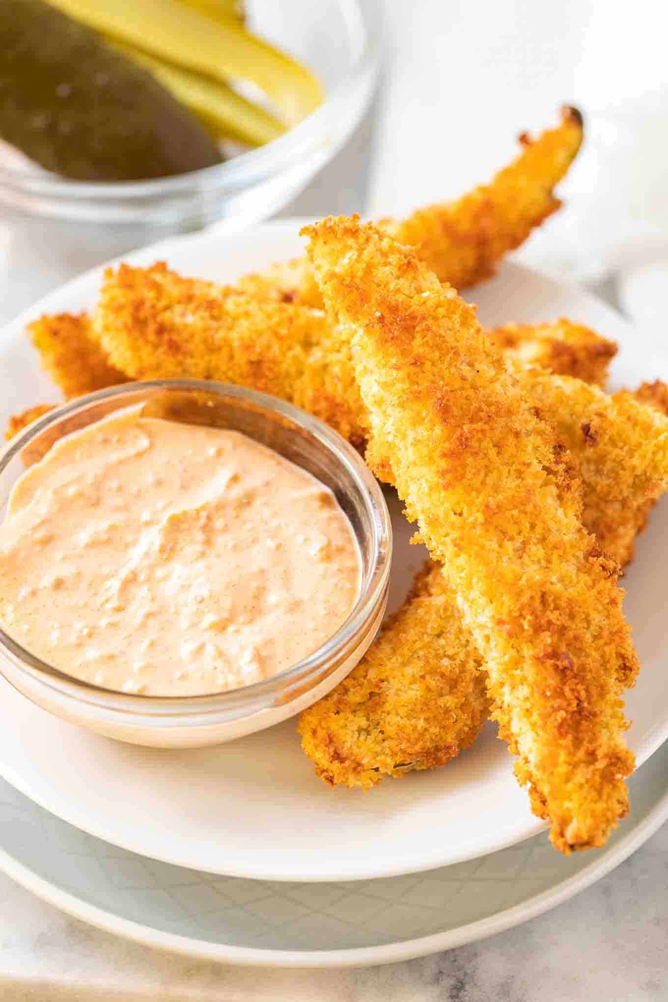 Une pile de cornichons à l'aneth frits à la friteuse à air sur une assiette blanche avec un petit bol en verre contenant une sauce.