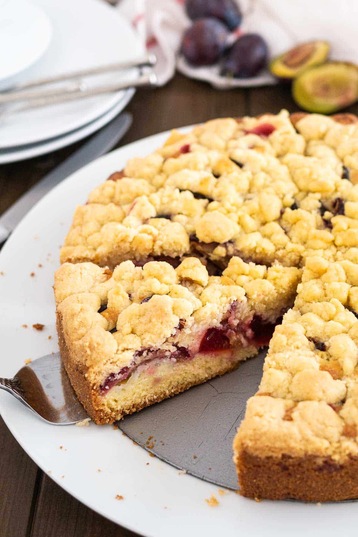 Un gâteau aux prunes avec streusel sur un fond de moule à charnière dans une assiette blanche. Un serveur à gâteau soulève une part.