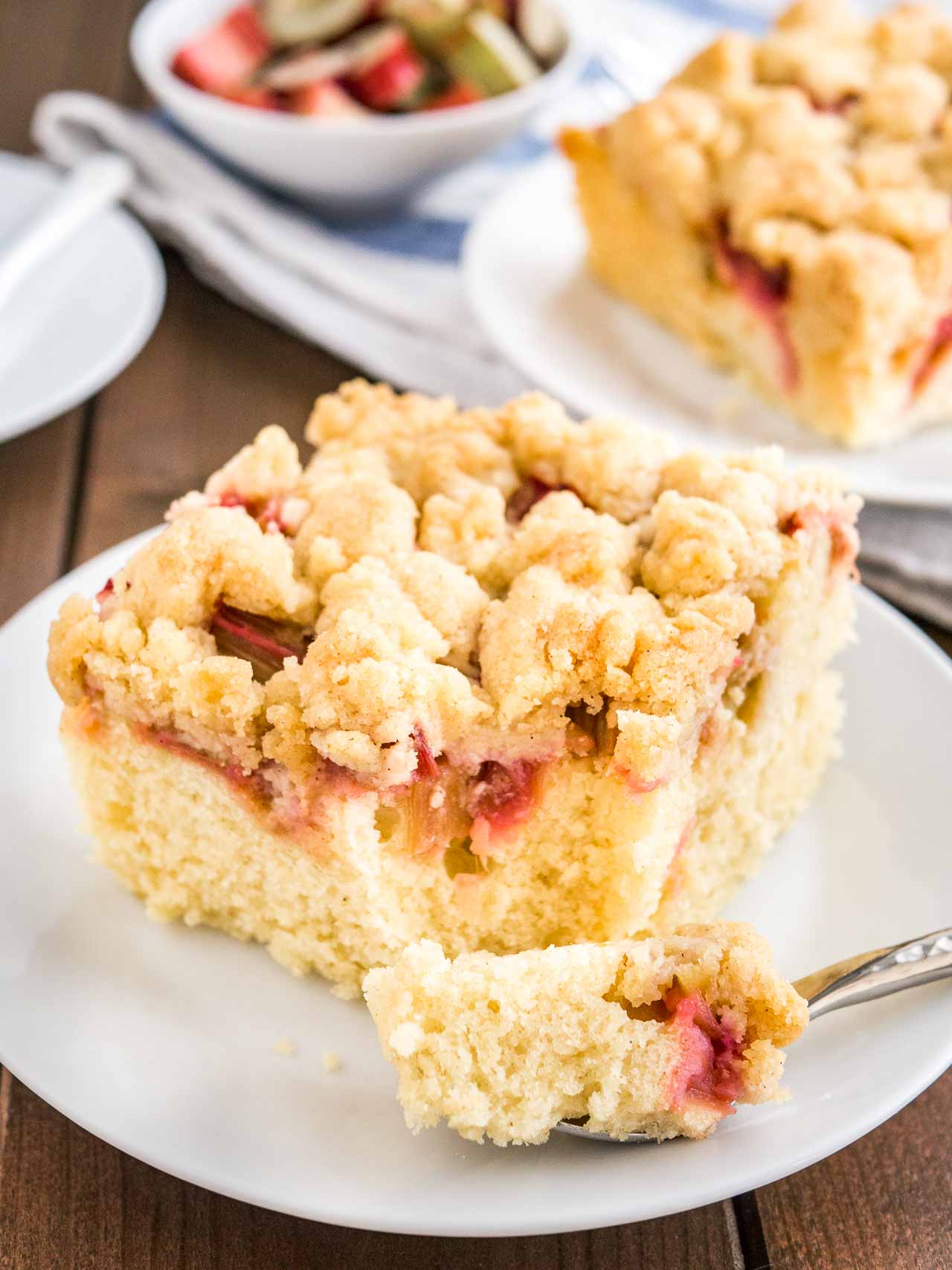 Gros plan d'une part de gâteau au café à la rhubarbe surmontée de streusel, sur une assiette blanche. Une fourchette a pris une part du gâteau. En arrière-plan, il y a une tasse de café et une autre part de gâteau sur une assiette.