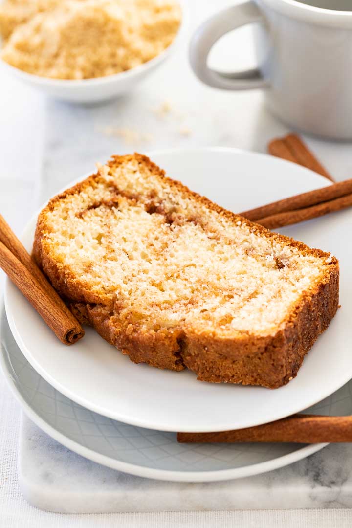 Une assiette blanche avec une tranche de pain au cannelle, garnie de quelques bâtons de cannelle et d'une tasse en métal grise avec un bol blanc contenant un mélange de sucre à la cannelle en arrière-plan.