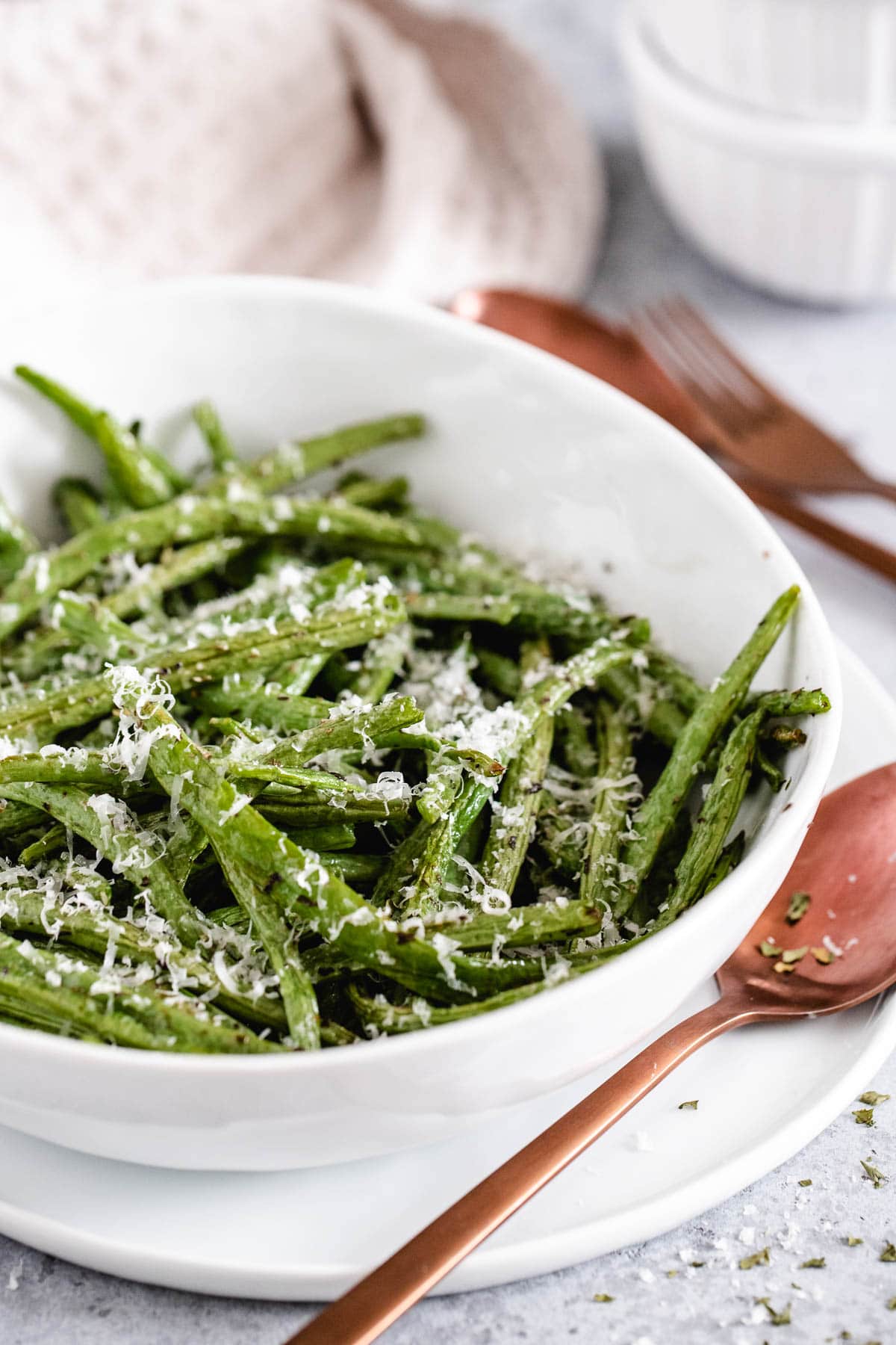 haricots verts cuits dans un plat blanc garnis de fromage râpé, à côté du bol se trouve une cuillère brune