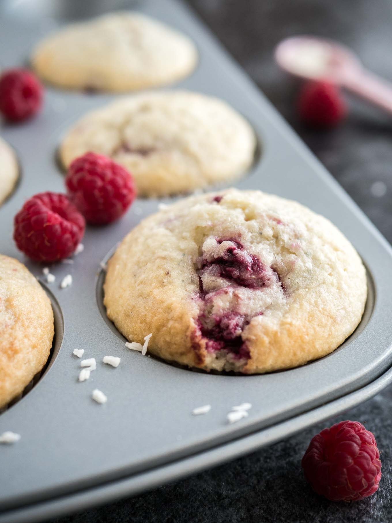 Gros plan d'un moule à muffins gris avec des muffins à la noix de coco et à la framboise garnis de framboises et de copeaux de noix de coco. Il y a des framboises et une cuillère à mesurer rose à côté.