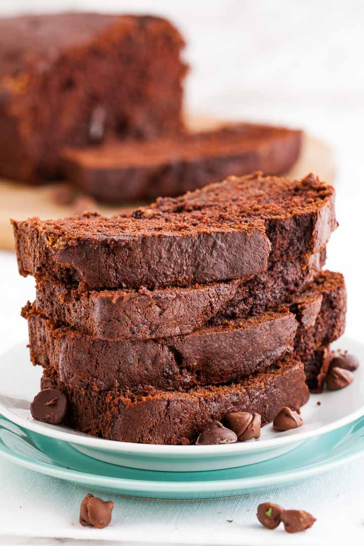 Une pile de 4 tranches de pain à la banane et au chocolat empilées sur une assiette blanche, garnie de quelques pépites de chocolat. Le pain entier est en arrière-plan.