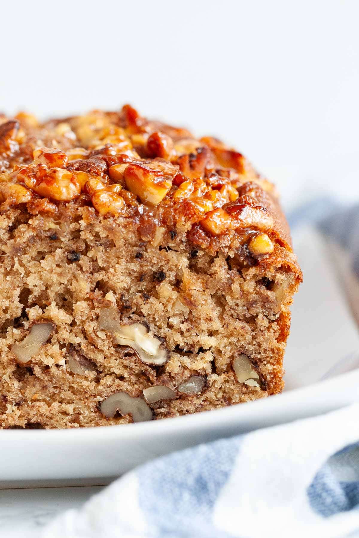 Ce pain à la banane est très moelleux et délicieux ! Surmonté de noix caramélisées et chargé de noix et de bananes écrasées, ma recette de pain à la banane est un favori de la famille qui est parfait pour le petit-déjeuner ou comme collation. Une façon idéale d'utiliser des bananes trop mûres ! #painbanane #painbananeauxnoix #recettesbananes #bananes #pain #recettespetitdéjeuner Gros plan d'un pain à la banane et aux noix avec une tranche coupée sur une assiette blanche rectangulaire à côté d'un torchon blanc et bleu.