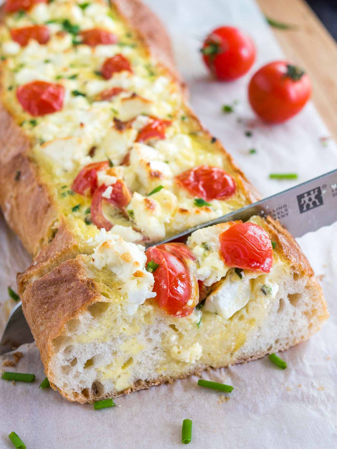 Un couteau à pain coupant du pain français fourré à la tomate et à la feta garni de tomates et de ciboulette sur une planche à découper en bambou recouverte de papier sulfurisé.