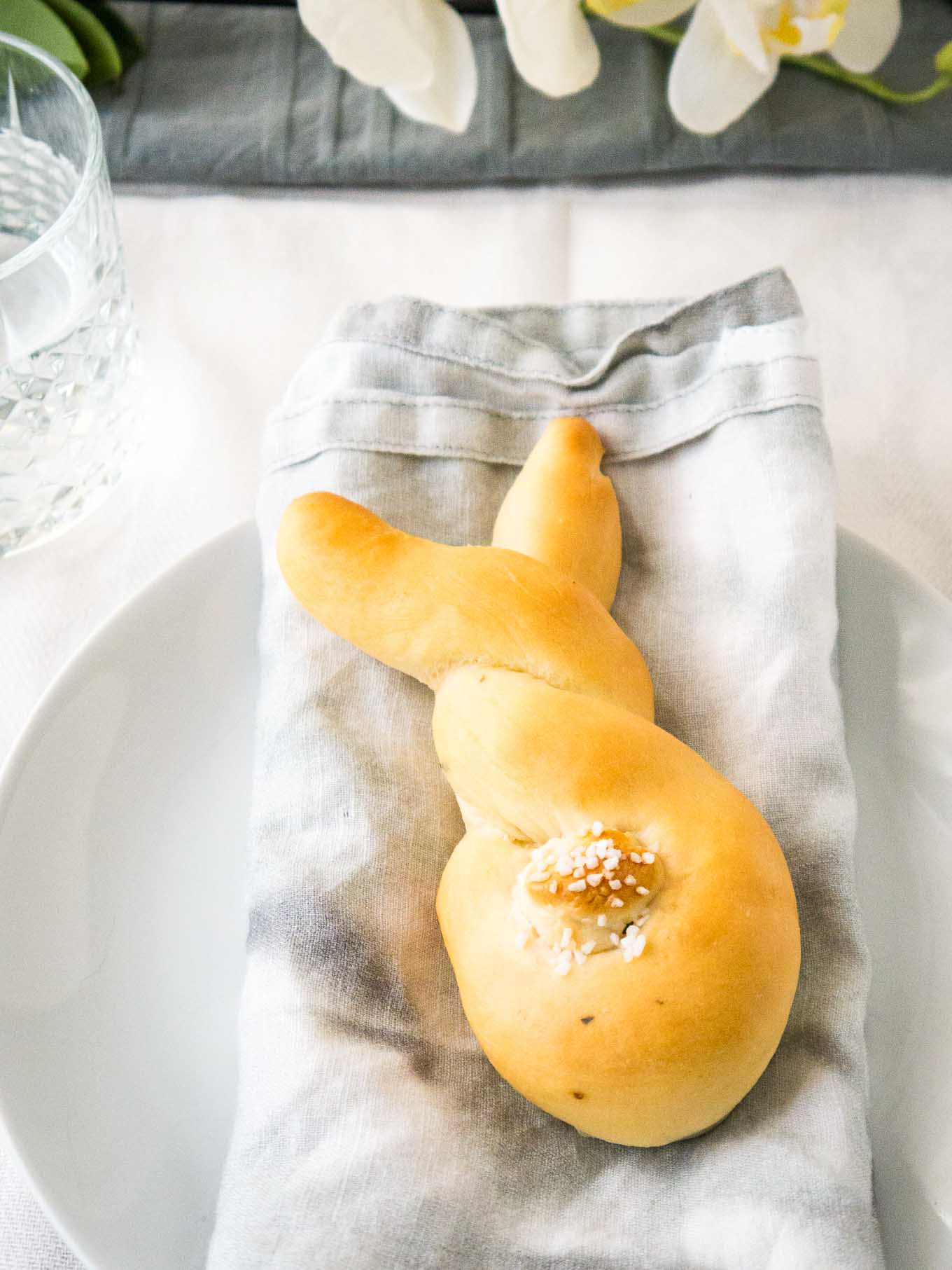 Pain en forme de lapin, avec une queue en pâte sur une serviette grise sur une assiette blanche à côté d'un verre d'eau.