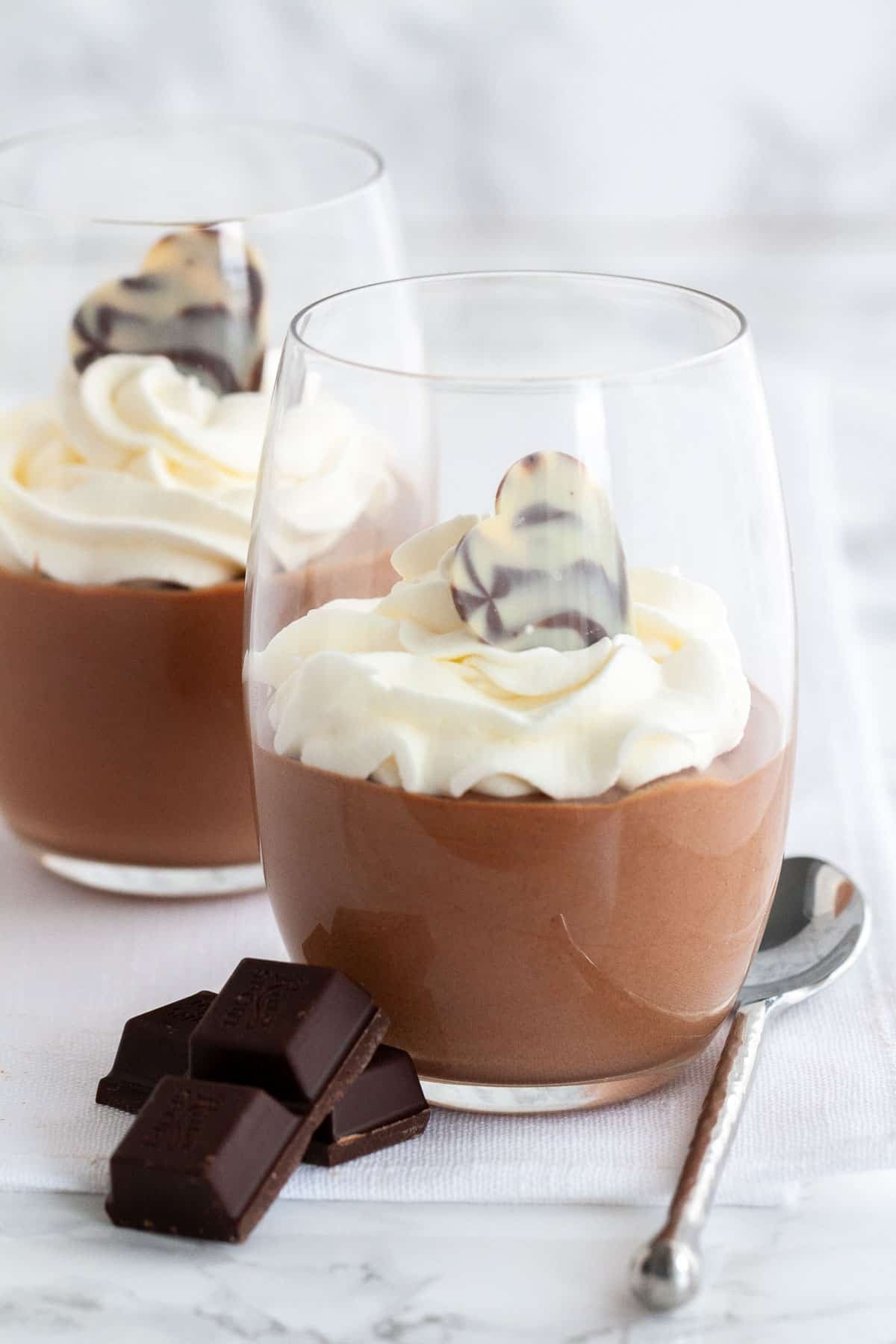 Cette recette de mousse au chocolat est super simple à réaliser maison et délicieusement légère et aérée. Une recette de mousse sans œuf qui est si aérienne et chocolatée et peut être appréciée par tout le monde. Assez élégante pour une fête, mais suffisamment facile à préparer chaque fois que vous avez une envie de chocolat ! Mousse au chocolat dans un verre, surmontée de crème fouettée à côté de morceaux de chocolat et d'une cuillère.