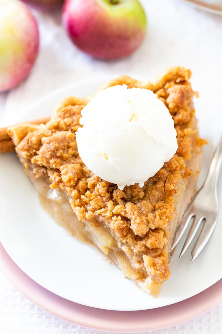 Une part de tarte aux pommes avec un streusel sur une assiette blanche empilée sur une assiette rose avec une fourchette, garnie de glace à la vanille, décorée de bâtons de cannelle et de pommes.
