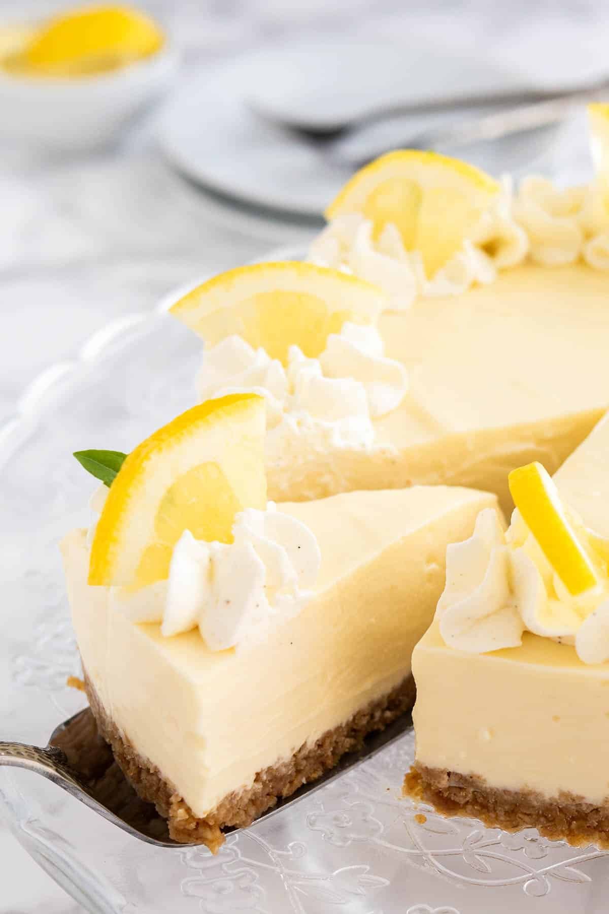 Une tarte à la crème au citron sur un plat en verre, garnie de crème fouettée et de quartiers de citron. Un serveur à gâteau soulève une part.