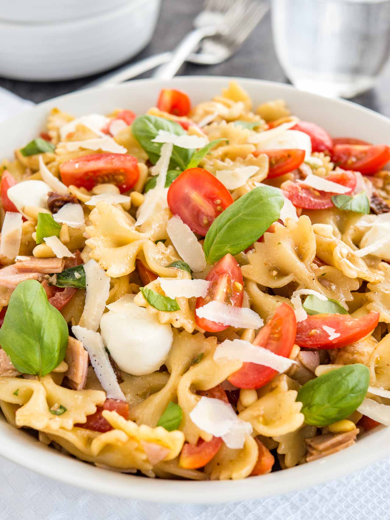 Gros plan sur un bol blanc contenant une salade de pâtes avec vinaigrette italienne, avec des tomates, de la mozzarella, des feuilles de basilic et des copeaux de parmesan. Le bol est posé sur un torchon blanc avec des fourchettes, un verre d'eau et des bols blancs.