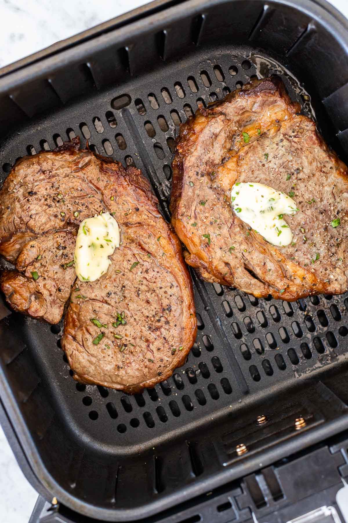 Deux steaks cuits avec un peu de beurre à l'ail sur le dessus dans un panier noir pour air fryer.