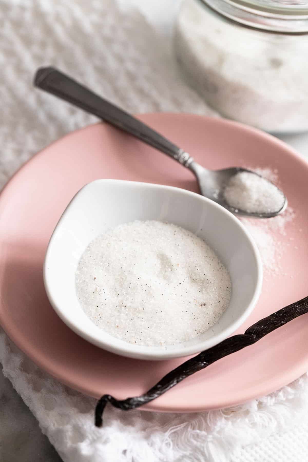 Un petit bol rempli de sucre vanillé sur une assiette rose avec une cuillère et une gousse de vanille à côté.