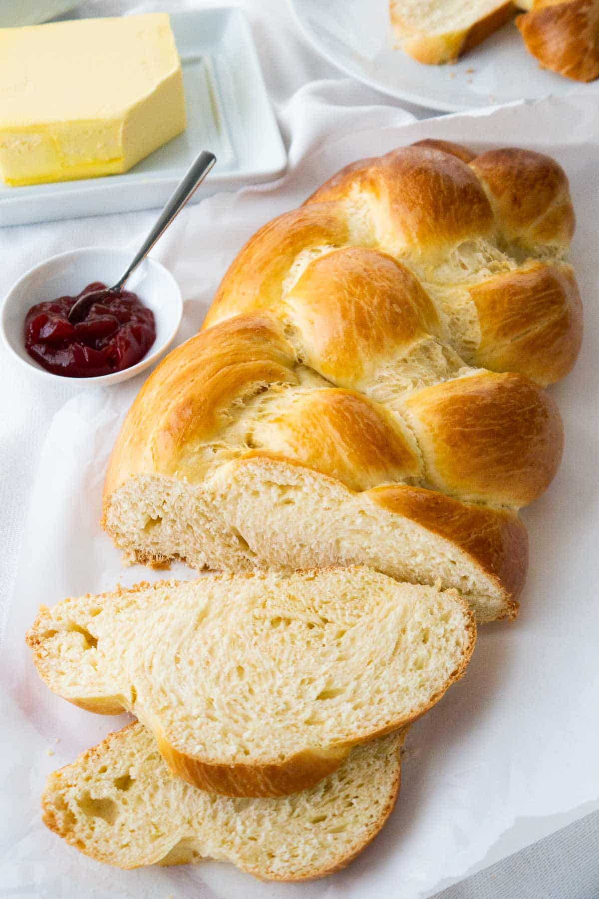 Le pain tressé est l'une de mes recettes préférées à préparer pour Pâques, mais il est délicieux tout au long de l'année ! Il est savoureux avec du beurre et de la confiture sur le dessus et les restes sont parfaits pour préparer du pain perdu ou du pudding au pain. Ce pain de levure tressé sucré facile est moelleux, légèrement sucré et enrichi en œufs et en crème épaisse pour une mie tendre ! #easter #easterbread #braidedbread #yeast #sweetbread #braidedloaf #osterzopf #hefezopf #braidedyeastbread #baking #easterbaking #easterrecipes Un pain sucré allemand, avec une tranche coupée et posée devant sur un torchon blanc à côté d'un petit bol blanc de confiture de fraises avec une cuillère.