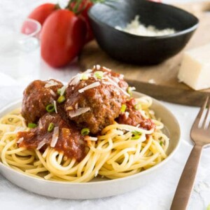 Une assiette grise avec des spaghetti et des boulettes de viande italiennes dans une sauce tomate sur un torchon blanc. Il y a une fourchette en bronze à côté, un verre d'eau, quelques tomates sur la vigne et une planche à découper en bois avec un bol noir de parmesan râpé en arrière-plan.