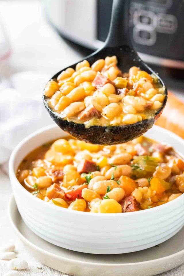 Un bol blanc contenant de la soupe de jambon et de haricots blancs, garni de persil. Une louche noire contenant un peu de la soupe est suspendue au-dessus du bol.