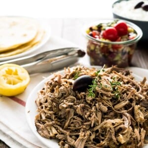 Une assiette de porc effiloché, garnie d'une olive et de branches de thym sur un torchon blanc avec une rayure rouge. Il y a un pot de salade grecque, un bol de tzatziki, une demi-lime, des pinces et une assiette de tortillas en arrière-plan.