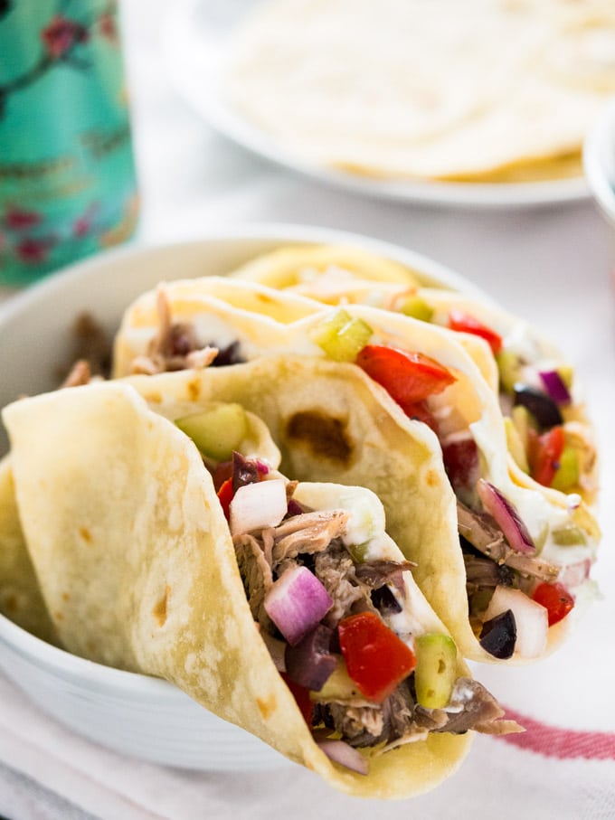 Un bol blanc de tacos à la grecque, garni de porc effiloché, de salade et de tzatziki. Il y a une assiette blanche de tortillas et une canette de thé glacé Arizona en arrière-plan.