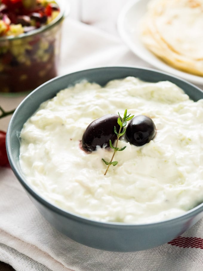 Un bol gris ardoise de Tzatziki, avec deux olives noires et un brin de thym sur un torchon blanc. Il y a une assiette blanche de tortillas et un bocal de salade grecque en arrière-plan.