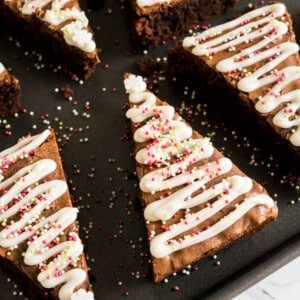 Une plaque de cuisson noire avec des brownies au pain d'épice en forme de triangle et des vagues de glaçage et de vermicelles pour les faire ressembler à de petits sapins de Noël.