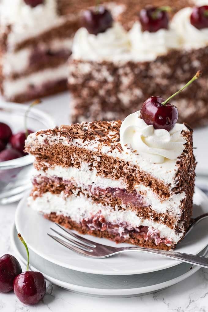 Un morceau de gâteau Forêt-Noire sur une assiette blanche à côté d'un couteau à gâteau. À l'arrière-plan se trouve un petit bol en verre rempli de cerises et un gâteau