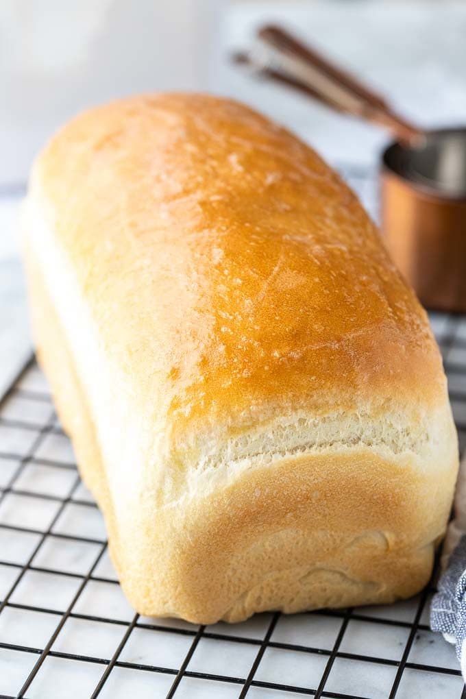 Une miche de pain blanc sur une grille de refroidissement noire à côté de quelques tasses à mesurer en bronze