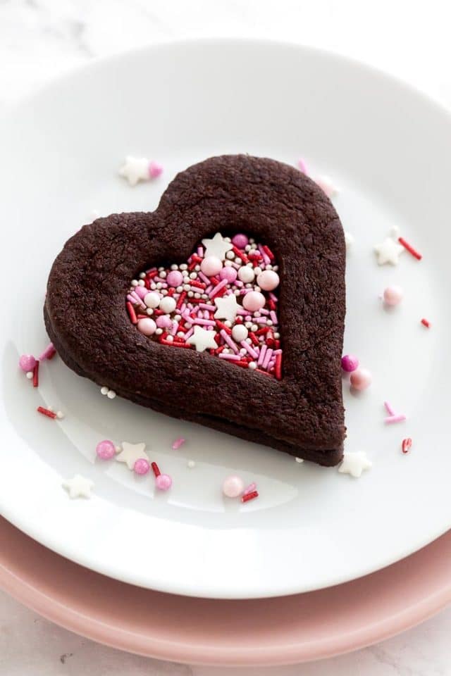 Un biscuit en chocolat en forme de cœur avec une indentation en forme de cœur remplie de vermicelles roses sur une assiette blanche