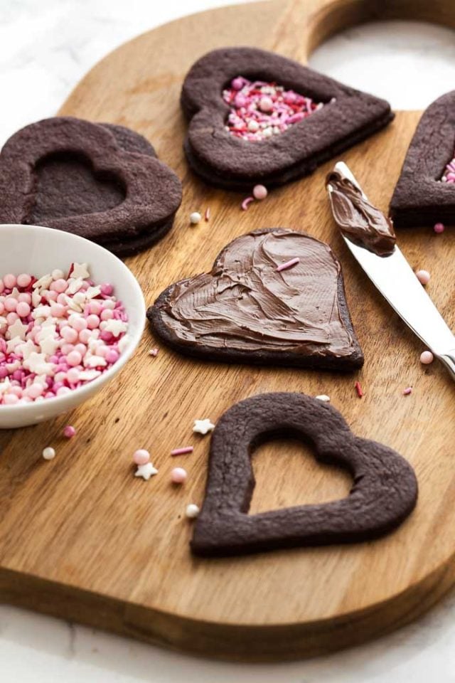 Une planche à découper en bois avec des biscuits au chocolat en forme de cœur à différents stades d'achèvement. Au premier plan, il y a la partie supérieure et inférieure du biscuit, un couteau a étalé du Nutella sur le bas. Le dessus a un petit cœur découpé. Il y a un petit bol de vermicelles à côté.