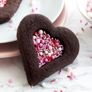 Un biscuit en chocolat en forme de cœur avec une indentation en forme de cœur remplie de vermicelles roses s'appuyant contre une pile d'assiettes garnies de vermicelles. Il y a un autre biscuit sur la pile d'assiettes et un petit bol blanc de vermicelles roses.