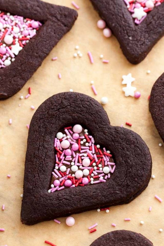 Vue du dessus de biscuits au chocolat en forme de cœur avec une indentation en forme de cœur remplie de vermicelles roses sur du papier sulfurisé avec des vermicelles à côté.