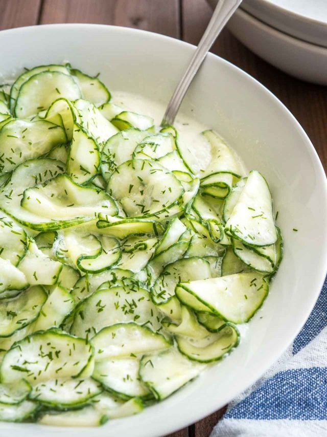 Gros plan de la salade de concombre allemande dans un bol de salade blanc avec une cuillère dedans sur un linge de cuisine blanc et bleu.