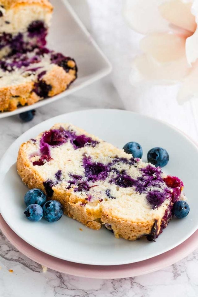 Une tranche de pain aux myrtilles sur une assiette blanche empilée sur une assiette rose, avec des myrtilles à côté d'une assiette rectangulaire avec une tranche de pain aux myrtilles dessus.