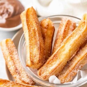 churros sucrés dans un bol en verre avec du papier sulfurisé, avec un petit bol blanc de nutella en arrière-plan
