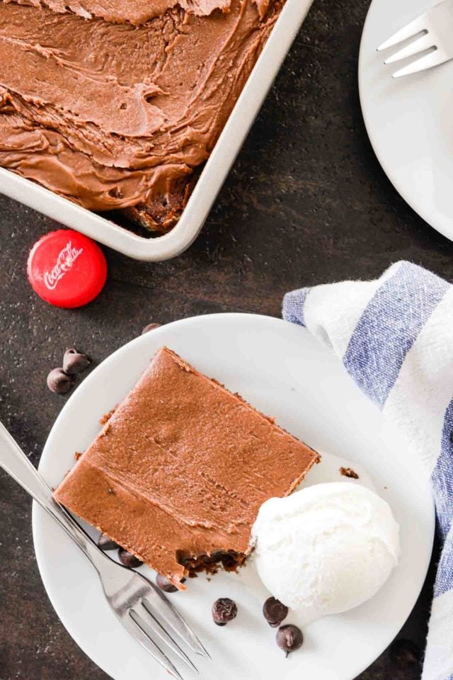 vue de dessus du gâteau au coca-cola sur une assiette blanche avec de la glace à la vanille, le coin d'un plat de cuisson avec le gâteau dedans et un bouchon de bouteille sur une table sombre.