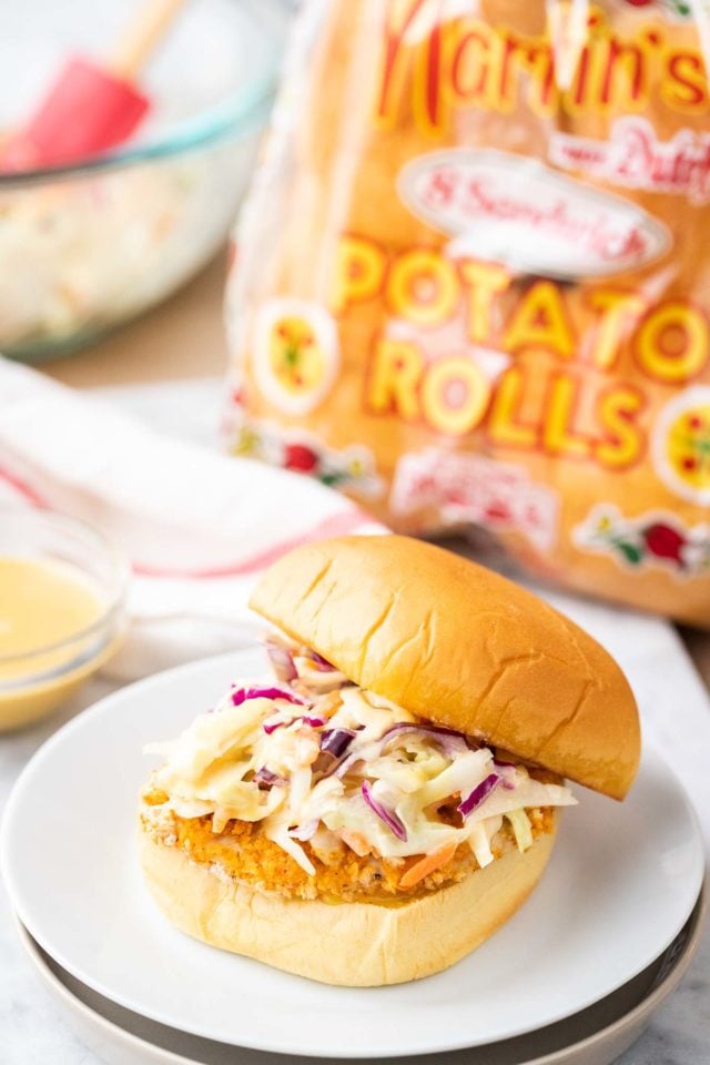 Un burger avec un pavé de poulet frit pané et de la salade de chou dont le dessus glisse sur une assiette blanche à côté d'un bol en verre avec une sauce, une serviette blanche et rouge, un bol en verre avec de la salade de chou et un sac de pains à hamburger
