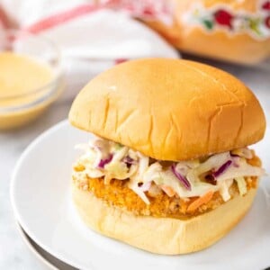Un burger avec un pavé de poulet frit pané et de la salade de chou sur une assiette blanche à côté d'un bol en verre avec une sauce, une serviette blanche et rouge et un sac de pains à hamburger
