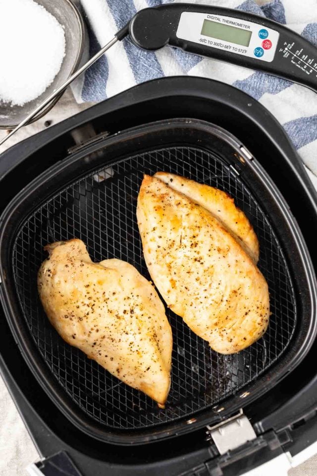 Deux poitrines de poulet frites dans un panier de friteuse à air sur un torchon blanc et bleu avec un bol sombre rempli de sel et un thermomètre à côté.