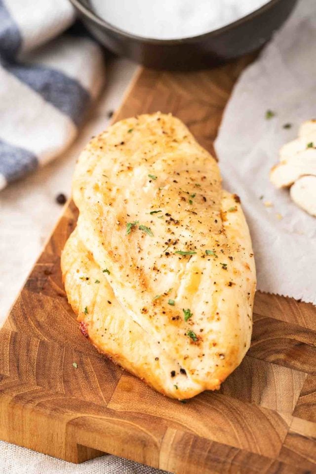Une poitrine de poulet grillée avec des herbes sur une planche à découper en bois sombre à côté de morceaux de poitrine de poulet tranchés et d'un torchon blanc et bleu.