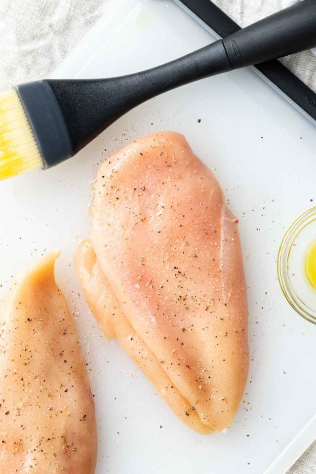 Deux poitrines de poulet crues sur une planche à découper en plastique, salées et poivrées. Un petit bol en verre avec de l'huile d'olive et un pinceau en silicone sont à côté.
